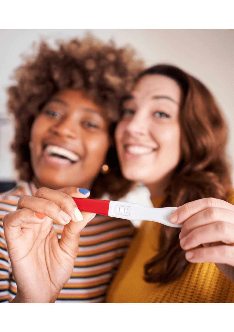 Two women, an interacial same sex couple, happily holding up a positive pregnancy test.