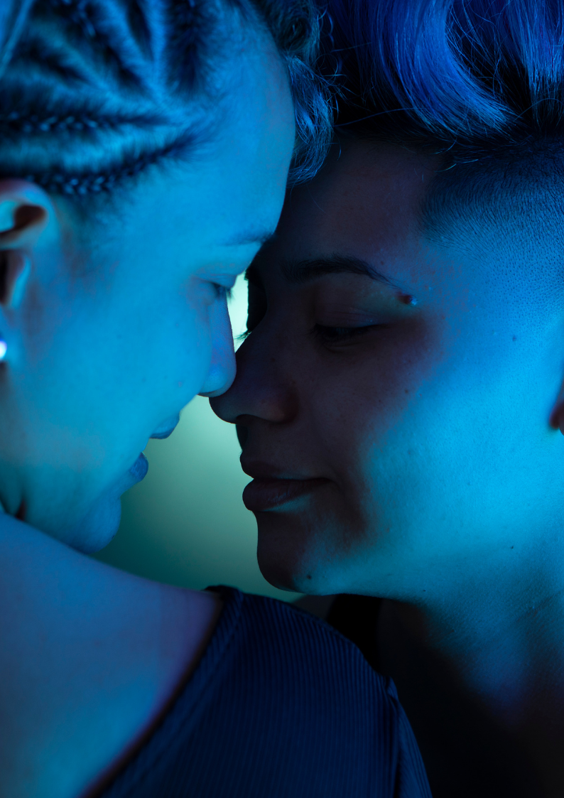 Two light-skinned, AFAB or female appearing people in a darkened blue lit setting with their faces almost touching, seeming about to kiss.