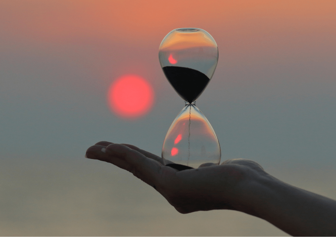 A feminine hand holds an hourglass palm up against a sunset with a red orb against blue grey horizon