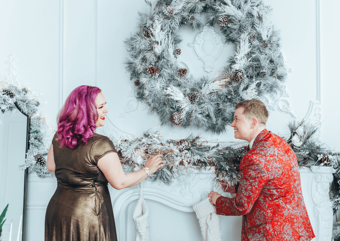 Two white queer people, one femme and one masc, wearing dress up clothes hang festive garlands in a winter holiday themed room.