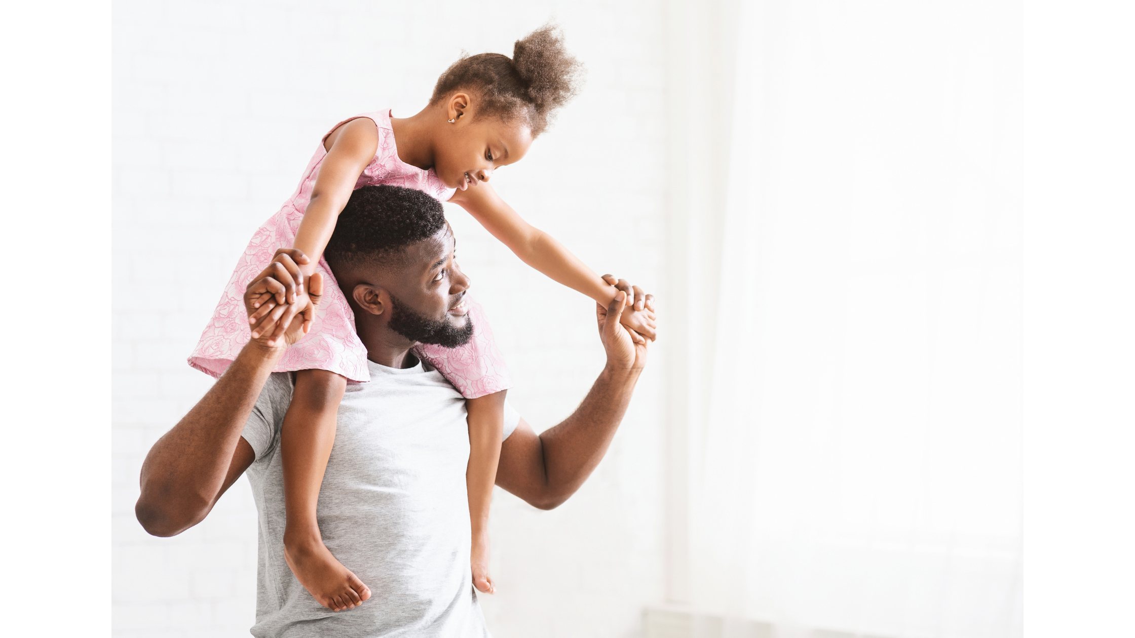 A black man carries a young black girl on his shoulders, holding both her hands and smiling up at her while she smiles down to him.